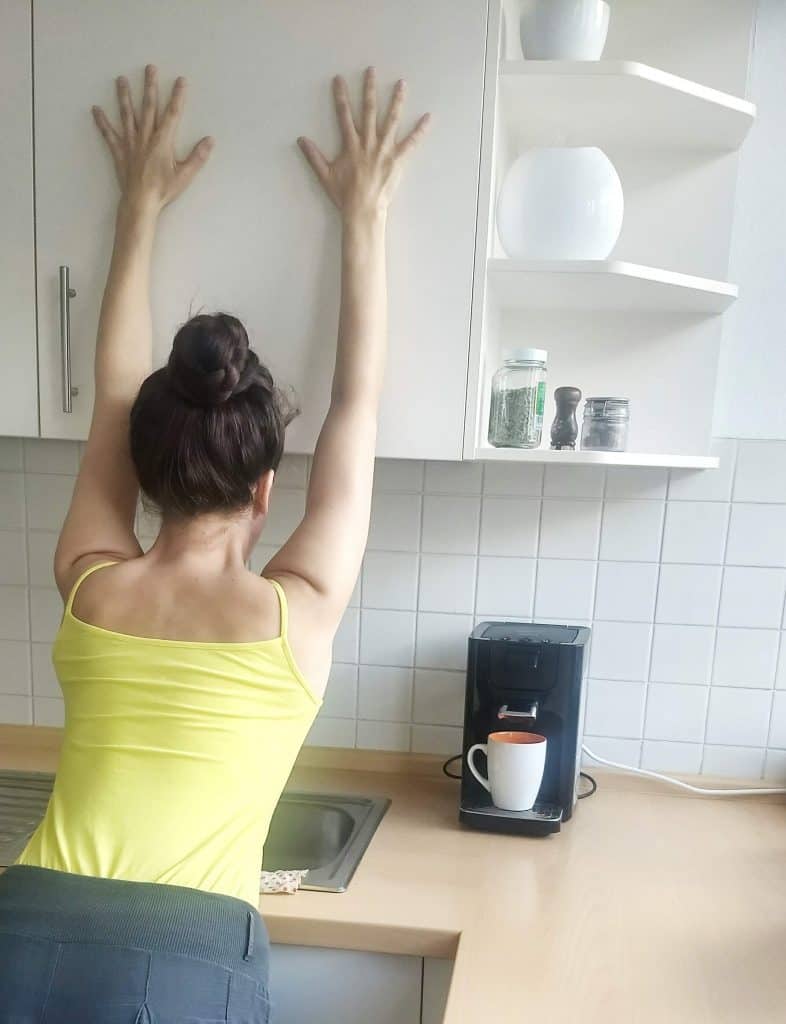 Image of a woman standing next to a coffee machine, stretching by reaching her arms up on the cupboard