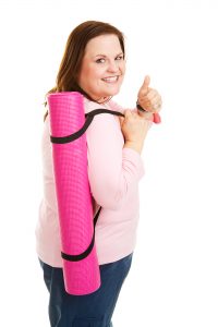 Image of a woman carrying a pink yoga mat, smiling at the camera and showing a thumbs up, ready for her beginners yoga on youtube