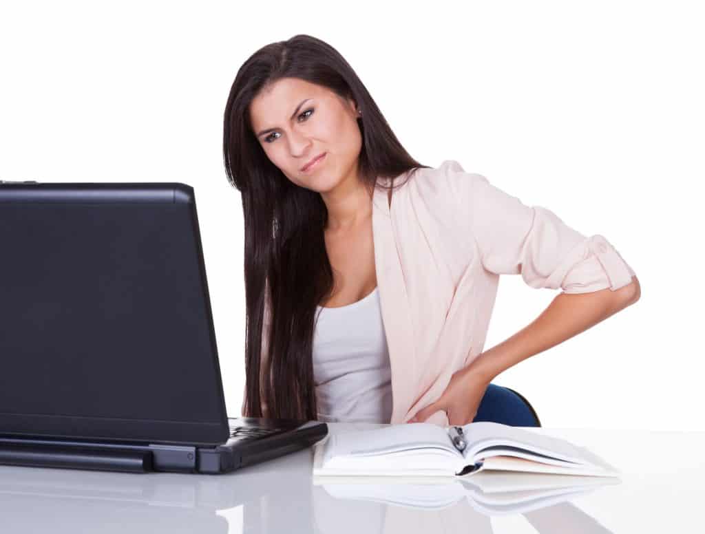 Image of a woman sitting in front of a laptop, holding her lower back and making a face of discomfort